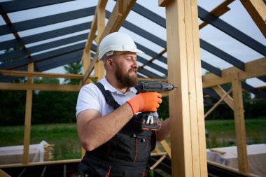 Ahşap çerçeve inşa ederken elektrikli tornavida kullanan inşaat işçisi. Koruyucu kasklı adam, eldiven. İnşaat, marangozluk, mühendislik ve inşaat süreci kavramı.