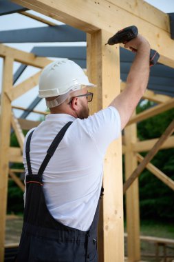 Ahşap çerçeve inşa ederken elektrikli tornavida kullanan inşaat işçisi. Koruyucu kasklı adam, eldiven. İnşaat, marangozluk, mühendislik ve inşaat süreci kavramı.