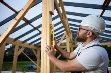 İnşaat işçisi, çerçeve alanındaki ahşap kirişin dikey hizasını kontrol etmek için ruh seviyesini kullanıyor. Kasketli ve güvenlik gözlüklü bir inşaat işçisi açık havada ahşap bir yapı üzerinde çalışıyor..