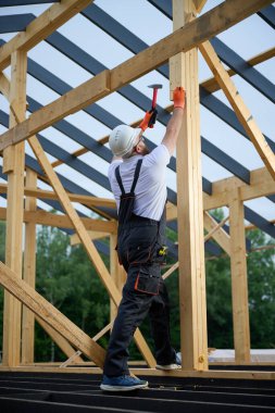 İnşaat işçisi inşaat alanındaki tahta kirişleri korumak için çekiç kullanıyor. Koruyucu kasklı, eldivenli ve ahşap iskelet üzerinde çalışan bir adam..