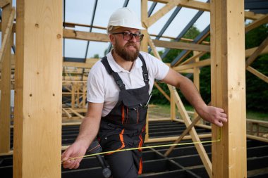 İnşaat işçisi inşaat iskeletinde tahta kirişi mezurayla ölçüyor. Kasklı bir bina, ahşap bina inşaatında çalışıyor..