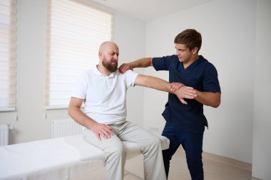 Kayropraktik uzmanı erkek hastaların omuzlarını klinikte düzeltiyor. Ağrıyı azaltmak, hareket kabiliyetini artırmak, yaralanmadan sonra rehabilitasyonu desteklemek ya da kas-iskelet bozukluğunu gidermek için fizyoterapist elle tedavi uyguluyor.