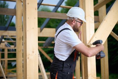 Ahşap çerçeve inşa ederken elektrikli tornavida kullanan inşaat işçisi. Koruyucu kasklı adam, eldiven. İnşaat, marangozluk, mühendislik ve inşaat süreci kavramı.
