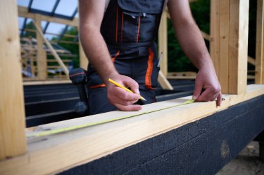 İnşaat alanında tahta kirişi kalem ve mezurayla işaretleyen inşaat işçisinin yakın çekimi. Ev iskeleti inşaatı sırasında hassasiyet ölçümü.