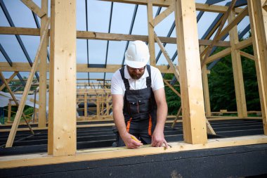 Construction worker marking wooden beam with pencil and tape measure at building site. Precision measurement during house frame construction.