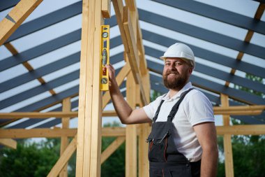 İnşaat işçisi, çerçeve alanındaki ahşap kirişin dikey hizasını kontrol etmek için ruh seviyesini kullanıyor. Kasketli ve güvenlik gözlüklü bir inşaat işçisi açık havada ahşap bir yapı üzerinde çalışıyor..