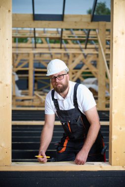 İnşaat işçisi inşaat iskeletinde tahta kirişi mezurayla ölçüyor. Kasklı bir bina, ahşap bina inşaatında çalışıyor..