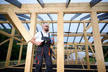 İnşaat işçisi ahşap ev iskeleti şantiyesinde mola verip su içiyor. İnşaatçı kaskı ve su şişesiyle dinleniyor..