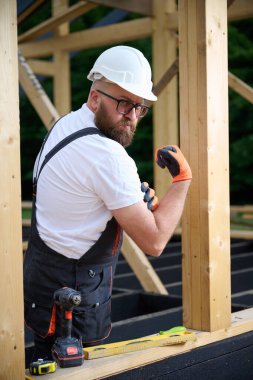 Eldiven ve tulumlu inşaat işçisi kolunu esnetiyor ve ahşap bir şantiyede gücünü gösteriyor. İş sırasında poz veren kendine güvenen bir zanaatkar. Güç, emek ve marangozluk kavramı.