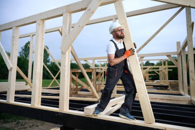 İnşaat işçisi ev iskeleti şantiyesine ahşap kiriş yerleştiriyor. Marangoz, yeni açık hava evi için ahşap yapı inşa ediyor..