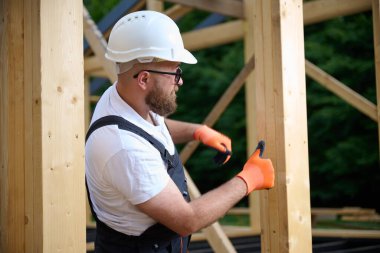 İnşaat işçisi, kasklı ve eldivenli. İnşaat alanında ahşap çerçeve ölçüyorlar. Kereste yapısı üzerinde çalışan odaklanmış erkek marangoz. El işi ve güvenlik kavramı.