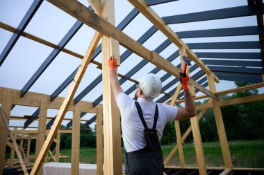 İnşaat işçisi inşaat alanındaki tahta kirişleri korumak için çekiç kullanıyor. Koruyucu kasklı, eldivenli ve ahşap iskelet üzerinde çalışan bir adam..