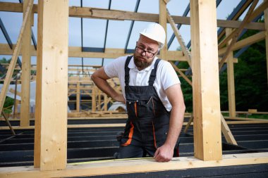 İnşaat işçisi inşaat iskeletinde tahta kirişi mezurayla ölçüyor. Kasklı bir bina, ahşap bina inşaatında çalışıyor..