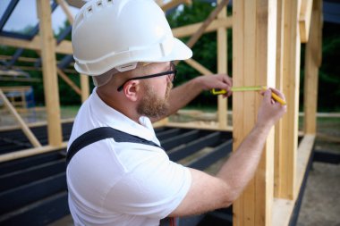 İnşaat işçisi inşaat iskeletinde tahta kirişi mezurayla ölçüyor. Kasklı bir bina, ahşap bina inşaatında çalışıyor..