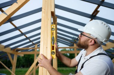 İnşaat işçisi, çerçeve alanındaki ahşap kirişin dikey hizasını kontrol etmek için ruh seviyesini kullanıyor. Kasketli ve güvenlik gözlüklü bir inşaat işçisi açık havada ahşap bir yapı üzerinde çalışıyor..