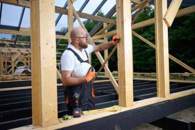 İnşaat işçisi kereste iskelet evindeki ahşap kiriş hizasını kontrol ediyor. Güvenlik teçhizatında miğfer tutan ve dışarıda ahşap yapı üzerinde çalışırken aletler kullanan bir bina..