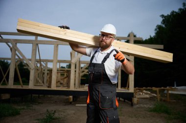 Construction worker carrying wooden beam on shoulder at building site. Carpenter in uniform and safety helmet working on timber house framework outdoors. Craftsmanship and construction work.
