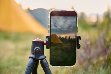 Bir tripodun üzerindeki akıllı telefon, sabahları dağlarda güneşin doğuşunun videosunu çeker. Dağlarda cep telefonu. Yalnız kamp. Cep telefonuyla dağlarda video yakalamak.