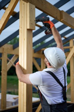 Ahşap çerçeve inşa ederken elektrikli tornavida kullanan inşaat işçisi. Koruyucu kasklı adam, eldiven. İnşaat, marangozluk, mühendislik ve inşaat süreci kavramı.