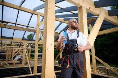 İnşaat işçisi ahşap ev iskeleti şantiyesinde mola verip su içiyor. İnşaatçı kaskı ve su şişesiyle dinleniyor..