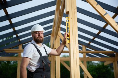 İnşaat işçisi, çerçeve alanındaki ahşap kirişin dikey hizasını kontrol etmek için ruh seviyesini kullanıyor. Kasketli ve güvenlik gözlüklü bir inşaat işçisi açık havada ahşap bir yapı üzerinde çalışıyor..