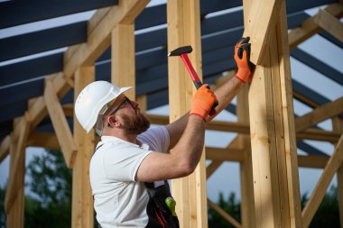 İnşaat işçisi inşaat alanındaki tahta kirişleri korumak için çekiç kullanıyor. Koruyucu kasklı, eldivenli ve ahşap iskelet üzerinde çalışan bir adam..