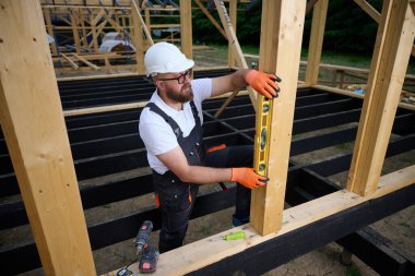 İnşaat işçisi, çerçeve alanındaki ahşap kirişin dikey hizasını kontrol etmek için ruh seviyesini kullanıyor. Kasketli ve güvenlik gözlüklü bir inşaat işçisi açık havada ahşap bir yapı üzerinde çalışıyor..