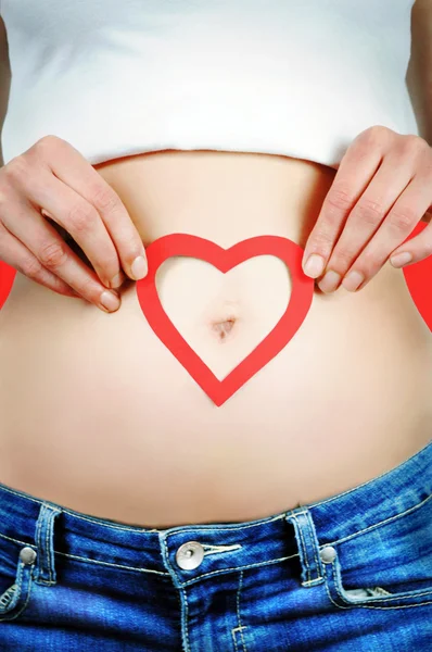 Nahaufnahme einer jungen Frau, die um ihren Bauchnabel herum eine rote Herzform auf dem Bauch trägt. roter Hintergrund. — Stockfoto