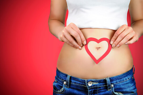 Close-up portrait of a young woman holding a red heart shape on her stomach around her navel. Red background.