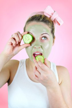 Beauty portrait of a funny woman wearing a pink headband, white top and a green facial mask is holding a slice of cucumber in front of her left eye and is about bite into another one. clipart