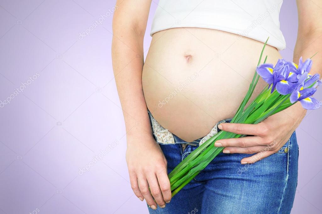 Close-up portrait of a bare womans baby bump and purple iris flowers on a purple background.