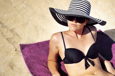 Sahilde bir havlu üzerinde siyah bikini, büyük siyah ve beyaz çizgili yaz şapka ve siyah güneş gözlüğü giyen güzel kadın istirahat.