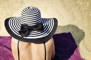Siyah bikini giyen zarif bir kadın ve büyük siyah beyaz çizgili bir yaz şapkası kumsaldaki havluda rahat bir şekilde yatıyor..
