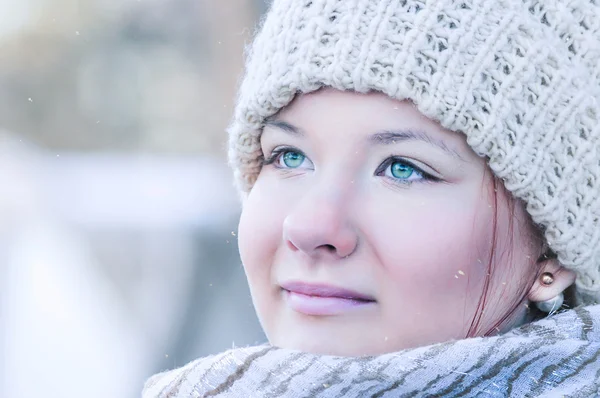 Menina bonita com olhos azuis brilhantes vestindo um chapéu de malha bege e um cachecol de pé fora em tempo frio em leve queda de neve . Imagens De Bancos De Imagens