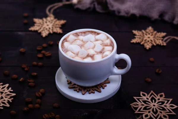 Cup of coffee with marshmallows and wooden snowflakes on dark wooden table. Winter concept, dark mood photo.