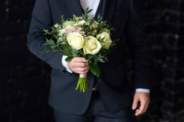 Takım elbiseli damat düğün buketini yakın planda tutuyor.