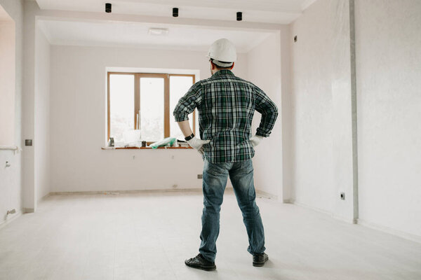 Ремонтник в каске стоит спиной в пустой комнате