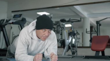 Bir adam spor paspası üzerinde tahta egzersizi yapıyor. Sweatshirt ve bere giyiyor. Spor aletleri arka planda görülebiliyor, fitness ortamını vurguluyor.