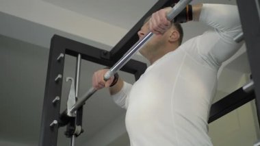 Bir adam spor salonu barfiks çubuğunda barfiks çekerek egzersiz yapıyor. Uzun kollu bir gömlek giyerken vücudunun üst kısmına odaklanıyor. Spor salonu eğitim için geniş bir alan sağlar..