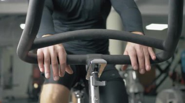 Spor salonundaki sabit bir bisikletin gidonunu tutan eller. Arka planda çeşitli spor aletleri var. Kişi bir antrenman seansında. Bisiklet eylemine odaklan.