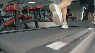 Spor salonundaki bir koşu bandında çalışan bir kişi, yakın çevrede durağan bisikletler ve ağırlık bankları da dahil olmak üzere çeşitli fitness ekipmanlarıyla kapalı bir alanda koşuyor..