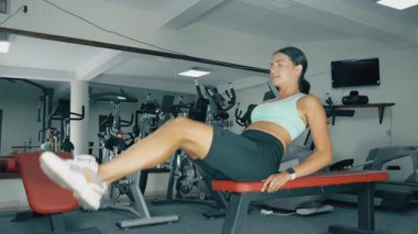 Bir kadın spor salonundaki bir bankta egzersiz yapıyor. Atletik giyinmiş ve vücut kaslarını hedef alan bir egzersiz sergiliyor. Teçhizat arka planda görünür.