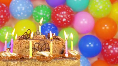 Mumları yanan bir doğum günü pastası masanın üzerinde duruyor. Pastanın arkasında çeşitli tonlarda renkli balonlar var. Atmosfer bir kutlamayı işaret ediyor..