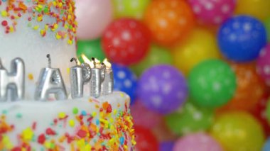 Renkli süslemelerle süslenmiş bir doğum günü pastasının üstünde dört mum var. Arka planda parlak renkli balonlar mevcuttur ve şenlikli bir sahne oluştururlar..