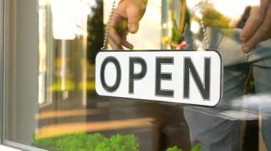 Dükkan vitrininde açık bir tabela asılı duruyor. İşletmenin müşterileri karşılamaya hazır olduğunu gösteriyor. Parlak gün ışığı girişi aydınlatıyor, yakınlardaki tesisleri aydınlatıyor..