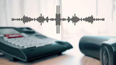 Ahşap bir masada dinlenen bir telefonun yakın çekimi. Ses dalgaları cihazın üzerinde belirir, sesi betimler. Arka plandaki perdelerin arasından ışık filtreleri.