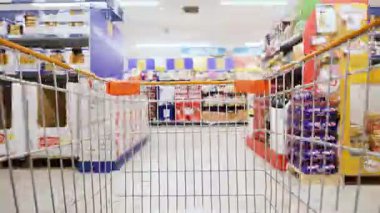 Önünde alışveriş arabası olan bir adam perakende mağazasında koridorda yürüyor. Raflarda çeşitli temizlik malzemeleri ve ev eşyaları var..