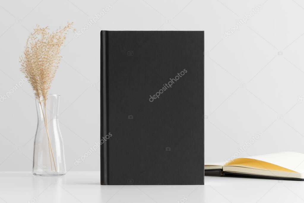 Black book mockup with a dried grass decoration and workspace accessories on a white table.