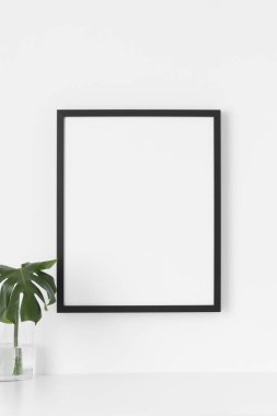 Duvarda siyah çerçeve, cam vazoda canavar yaprağı var..