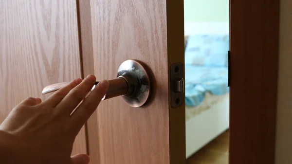 hand opening a wooden inner door and a bright bedroom with a white bed and a blue bedspread visible through the opening, female fingers pressing on the metal handle of the entrance room door